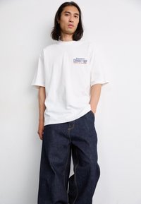 White short-sleeve t-shirt with a graphic logo on the chest, paired with loose dark blue denim jeans featuring yellow stitching.