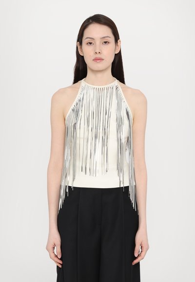 Haut halter avec des franges argentées et un corsage blanc ajusté, présentant des cordons métalliques qui tombent en cascade, associé à un pantalon noir à jambes larges.