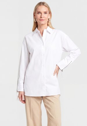 Blonde woman wearing a white long-sleeve button-up shirt with zipper cuffs and beige pants, standing with one hand on her hip.