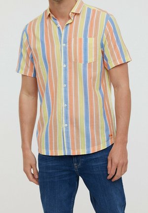 Męska koszula na krótkie rękawy zapinana na guziki z pionowymi pastelowymi paskami w kolorze żółtym, niebieskim, pomarańczowym i białym, zestawiona z ciemnoniebieskimi dżinsami.