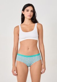Donna in piedi di fronte, indossa un reggiseno sportivo bianco e mutandine turchesi con un motivo floreale rosa, su uno sfondo semplice.