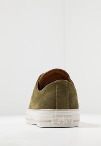 Sneaker verde in suede con suola in gomma bianca, con accenti marroni e un logo sul tallone. Design minimalista, senza lacci, dal profilo basso.