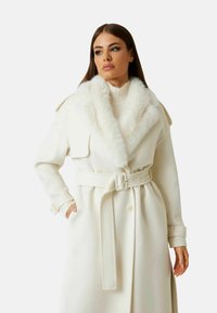 Cappotto bianco in lana con colletto in pelliccia ecologica, vita con cintura, tasche laterali, maniche lunghe e sottili dettagli con bottoni. Texture liscia e design caldo.