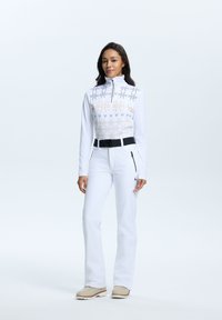 Top blanco de manga larga con media cremallera y pecho estampado, acompañado de pantalones blancos ajustados, con un cinturón negro y un bolsillo con cremallera.