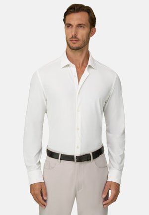Homme portant une chemise blanche à manches longues boutonnée et un pantalon beige clair avec une ceinture noire, debout devant un fond uni.