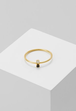 Bague fine en or avec deux petites pierres carrées, une claire et une noire, présentée sur un socle blanc sur un fond gris.