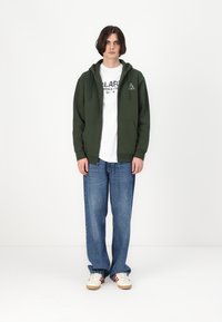 Grønn zip-up hettejakke med en frontlomme, over en hvit grafisk t-skjorte, kombinert med vide blå jeans og hvite joggesko.