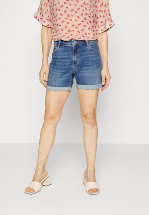 Kvinde iført optrukne blå denimshorts, en pink blomsterprintet bluse og beige åbne blokhæls-sandaler står på en ensfarvet hvid gulv.