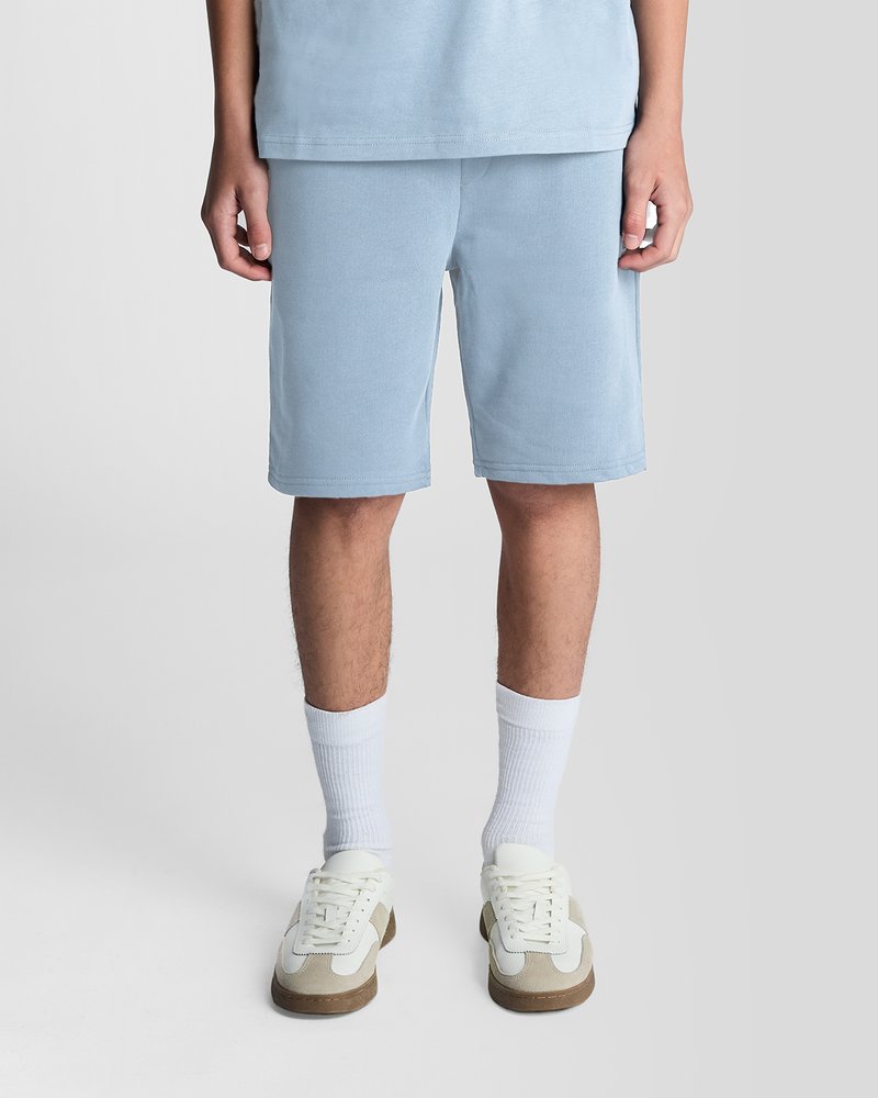 Persona che indossa pantaloncini azzurri chiari, calzini bianchi e sneakers bianche con dettagli beige, in piedi davanti a uno sfondo grigio uniforme.