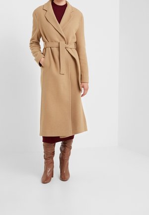 Femme portant un manteau camel ceinturé par-dessus une robe bordeaux avec des bottes marron à talons montant aux genoux, debout devant un fond gris uni.