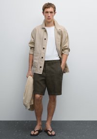 Beige linen shirt jacket, white t-shirt, dark green shorts, and flip-flops. Sweater held casually. Clean lines, minimalistic style.