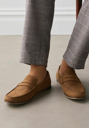 Braune Wildleder-Loafer getragen mit grauen karierten Hosen, dargestellt auf einem hellen Boden vor einem weißen Wandhintergrund.