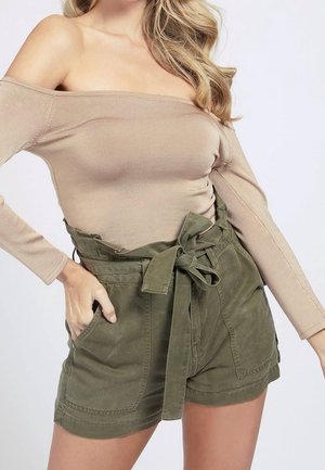 Femme portant un haut beige à manches longues et épaules dénudées ainsi qu'un short paperbag taille haute vert olive avec un lien devant et des poches.