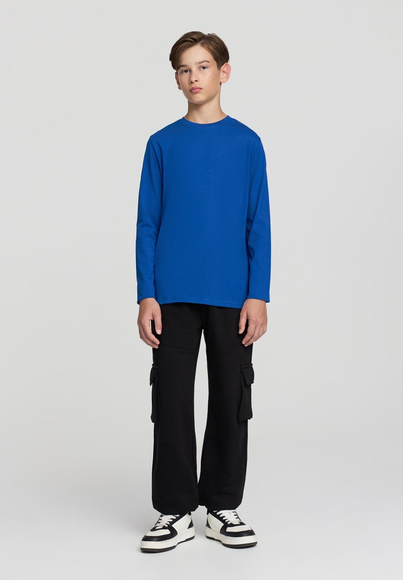 Niebieska koszula z długim rękawem z miękkiego materiału, zestawiona z czarnymi spodniami cargo z bocznymi kieszeniami. Obuwie to czarno-białe sneakersy.