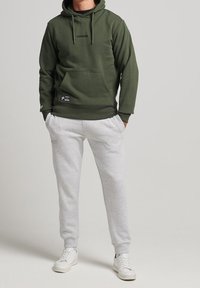 Olivgrön hoodie med en framficka och snörning, tillsammans med ljusgrå träningsbyxor. Vita sneakers fulländar looken.