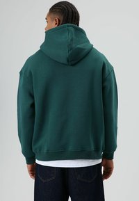 Groene hoodie met een losse pasvorm, geribbelde manchetten en zoom. Gladde stoftextuur, minimalistisch ontwerp, gedragen over een wit shirt en donkere spijkerbroek.