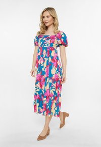 Flieder maxi Kleid in Blau mit pink und gelben Mustern, Puffärmeln, einem quadratischen Ausschnitt und einem gestuften Saum. Braune Absatzschuhe runden den Look ab.
