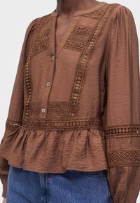 Blouse marron à manches longues avec panneaux en dentelle, devant boutonné, bas péplum et épaules bouffantes, portée avec un jean en denim bleu.