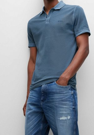 Blaues Polohemd aus strukturiertem Stoff mit einer drei-Knopf-Leiste und dezentem Logodetail, kombiniert mit hellblauen, abgenutzten Jeans.