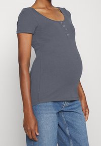 Mujer embarazada vistiendo una camiseta de manga corta de canalé gris con botones y unos jeans de mezclilla azul frente a un fondo liso.