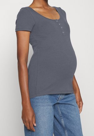 Mujer embarazada vistiendo una camiseta de manga corta de canalé gris con botones y unos jeans de mezclilla azul frente a un fondo liso.