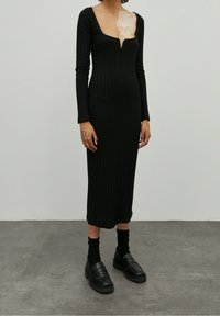 Schwarzes, geripptes Midikleid mit langen Ärmeln und tiefem, quadratischem Ausschnitt, kombiniert mit schwarzen Slippern und gerippten Socken, vor einem neutralen Hintergrund präsentiert.