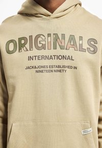 Beige hoodie with a front pocket, featuring bold "ORIGINALS" text in green and camouflage patterns, and smaller branding underneath.
