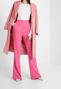 Cappotto di lana rosa sopra un maglione bianco a righe e pantaloni larghi rosa acceso. Scarpe a punta rosa chiaro completano l'outfit.