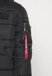 Sort pufferjakke med en kviltet design, lynlås lomme på ærmet og et rødt stofmærke med teksten "Fjern før flyvning."