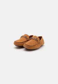 Brune ruskind moccasinsko med rundt tå, dekorativ syning og en metallaccent foran. Lavprofil design.