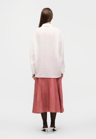 Blouse blanche texturée à manches longues, associée à une jupe midi en daim rose et des collants noirs, le tout complété par des chaussures à talons blanches.