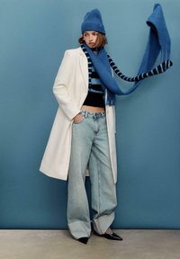 Cappotto bianco, maglione nero corto, jeans azzurri a gamba larga, berretto blu e lunga sciarpa blu a righe con frange, in piedi davanti a uno sfondo blu.