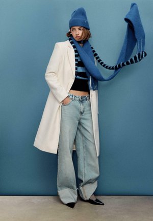 Beli plašč, črn skrajšan pulover, svetlo modre široke kavbojke, modra kapa in dolg modro črtast šal z resicami, stoji pred modrim ozadjem.