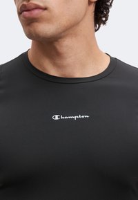 Κοντινό πλάνο ενός ατόμου που φοράει μαύρο t-shirt με στρογγυλή λαιμόκοψη της μάρκας Champion, που δείχνει τη λαιμόκοψη και το λογότυπο στο στήθος.
