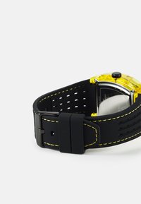 Schwarze Gummiuhr mit gelben Nähten, quadratischem Gehäuse und transparentem gelben Lünette. Verfügt über ein perforiertes Armband und eine Metallschließe.