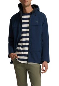 Chaqueta con capucha navy de textura suave, con cremallera y dos bolsillos. Lleva puesta una camisa de rayas en tonos navy y blanco. Pantalones verde oliva.