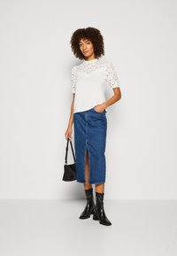 Hvid top med broderet øjelet-design og korte ærmer, parret med en blå denimnederdel med en slid foran, sorte ankelstøvler og en lille taske.