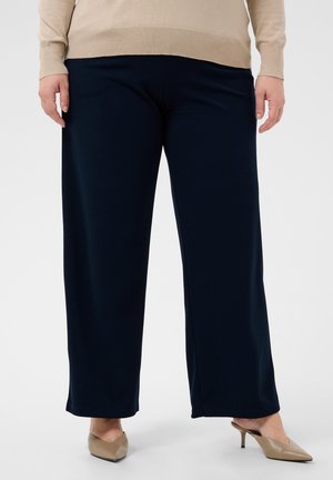 Personne portant un pantalon noir à jambes larges, un haut beige à manches longues et des escarpins beiges à bout pointu, debout sur fond blanc.