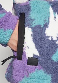 Fleecová bunda v purpurovej, tyrkysovej a bielej farbe s abstraktným vzorom. Obsahuje čierny zips na vrecku a štruktúrovaný, jemný povrch.