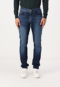 Jeans din denim de culoare albastru închis, cu un croi drept, având cinci buzunare, închidere cu nasture și ușoară decolorare. Purtați cu adidași albi.