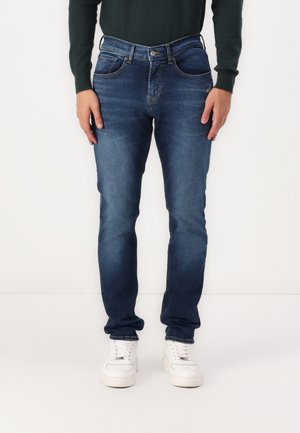 Mørkeblå denimjeans med rette ben, som har fem lommer, mønsterknap og subtil fading. Brukt med hvite sneakers.
