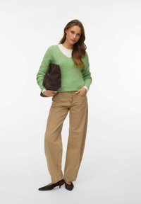 Femme portant un pull vert, un pantalon large beige et des talons noirs, tenant un grand sac à main en daim brun foncé sur un fond blanc.