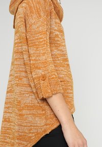 Side view of a person wearing a marled orange knit sweater with elbow-length sleeves rolled and buttoned up.