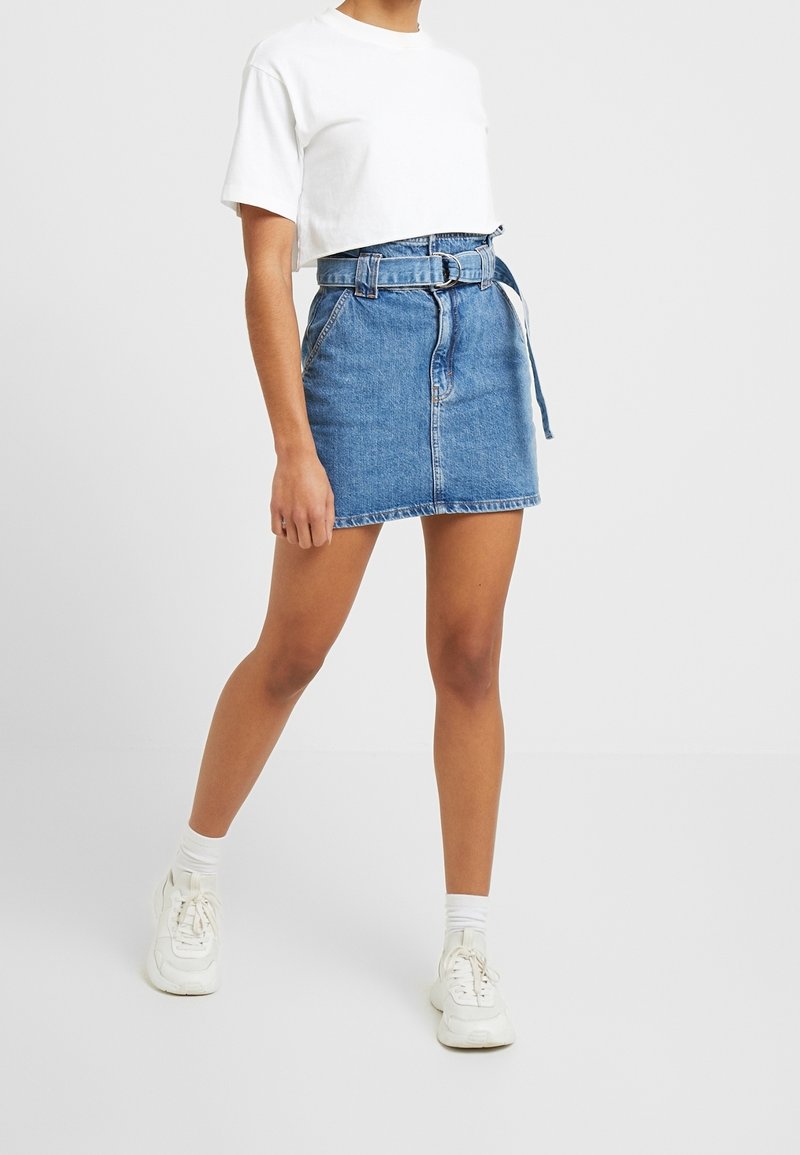 Sininen farkku-minihelma, jossa on vyöllinen vyötärö, edessä vetoketju ja taskut. Yhdistetty valkoiseen lyhythihaiseen t-paitaan ja valkoisiin lenkkareihin.