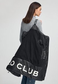 Svart vadderad jacka med fleecetyg på insidan, stor vit text "CLUB" vid fållen, quiltad textur, justerbara dragsnören och synliga skötselrådsetiketter.