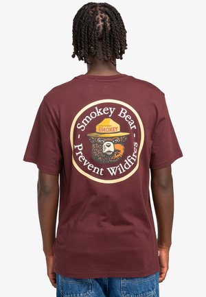 Camiseta de algodón burdeos con un gran gráfico circular de Smokey Bear y el texto "Previene incendios forestales" debajo. Manga corta y ajuste relajado.