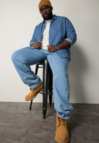 Blå denimskjorta, vit t-shirt, ljusblå jeans och bruna stövlar. Sitter på en svart pall med en texturerad grå golvbakgrund.