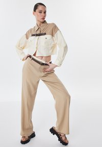 Camicia a maniche lunghe beige e crema, tagliata, con due tasche sul petto e dettagli di cintura marrone, abbinata a pantaloni larghi beige e sandali neri.