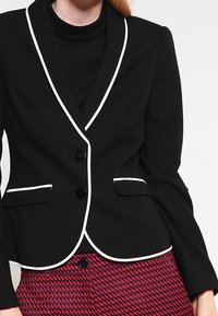 Svart kavaj med vit piping, knappstängning med en knapp, två framfickor och en strukturerad passform. Tillsammans med en mörk polotröja.