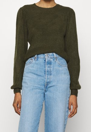 Person trägt einen olivgrünen Pullover mit langen Ärmeln, der in eine hoch taillierte hellblaue Jeans gesteckt ist, steht vor weißem Hintergrund.